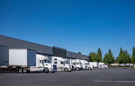 Different industrial grade for national wide freights big rig semi trucks with semi trailers standing at row at warehouse dock gates loading cargo for the next long haul and local deliveriesの写真素材