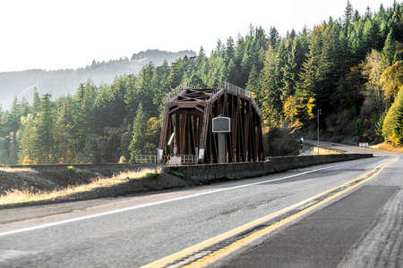Narrow truss railroad bridge across the inflow in the Columbia River floodplain along the highway road with high mountains and cliffs on the banks overgrown with autumn trees in Columbia Gorgeの写真素材