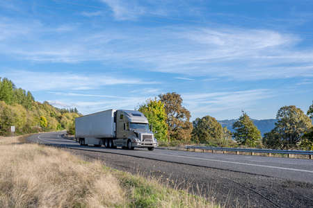 Industrial big rig gray semi truck tractor transporting commercial cargo in refrigerator semi trailer driving on the narrow winding highway road with autumn forest on the sides in Columbia Gorgeの写真素材
