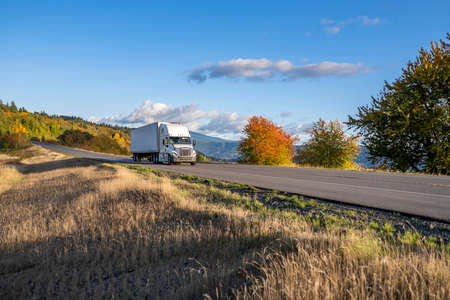 Industrial big rig white semi truck transporting commercial cargo in dry van semi trailer driving on the narrow highway road at spectacular Columbia River Gorge with yellow trees and grassの写真素材