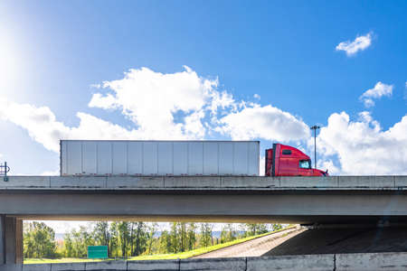 Industrial powerful red big rig classic semi truck transporting commercial cargo in dry van semi trailer running on the concrete bridge across the wide highway road on the cloud sky backgroundの写真素材