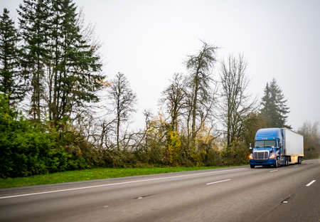 Industrial powerful blue big rig semi truck with turned on headlights transporting commercial cargo in dry van semi trailer driving on the twilight evening highway road for timely deliveryの写真素材