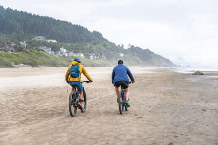 Two amateur men in in warm clothes travel side by side on the mountain bicycles along the foggy Northwest Pacific Ocean preferring an active healthy lifestyle tempering the body in any weatherの写真素材