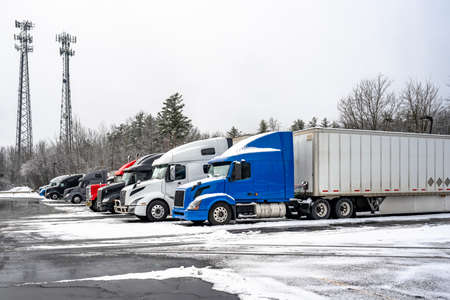 Big rigs semi trucks with different semi trailers standing for truck driver rest and continue the route according to the log-book on the winter truck stop parking lot with snow and ice surfaceの写真素材