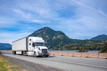 Clean well maintained Big rig white industrial grade powerful semi truck tractor with frozen cargo in refrigerator semi trailer driving on the road along the Columbia River in Columbia Gorge areaの写真素材