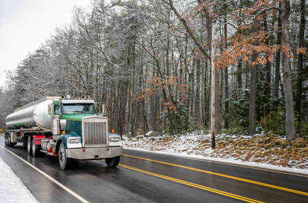 Industrial green day cab classic bonnet big rig semi truck transporting liquid cargo in long oval cistern tank semi trailer running on the wet winter road with snow on the sides and snowy forestの写真素材