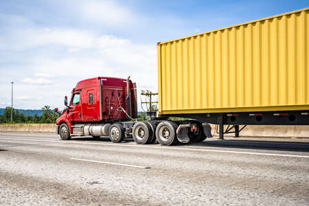 Low cab profile dark red big rig bonnet industrial semi truck tractor transporting commercial cargo in container semi trailer driving for delivery on the wide multiline straight highway roadの写真素材