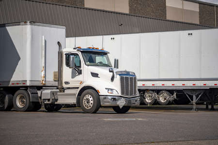 Big rig day cab white semi truck with dry van semi trailer in row with another semi trailers standing for unload commercial cargo in warehouse dock with gates on the industrial parking lotの写真素材