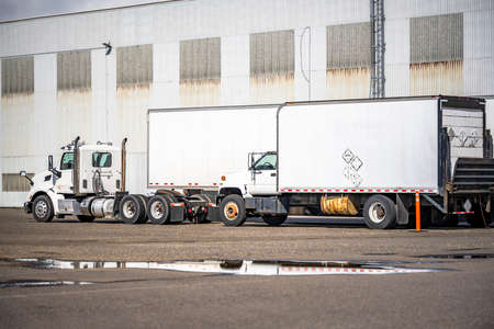 Big rig day cab semi truck with dry van semi trailer and small rig truck with box trailer standing on the industrial parking lot beside warehouse building waiting for the next load for next freightの写真素材