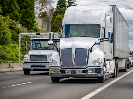 Loaded classic big rig white semi truck tractor transporting commercial cargo in dry van semi trailer running side by side with small rig semi truck on the wide multiline highway roadの写真素材