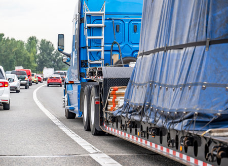 Industrial High cab blue big rig long haul semi truck transporting covered and fastened commercial cargo on step down semi trailer driving on the multiline highway road with another trafficの写真素材