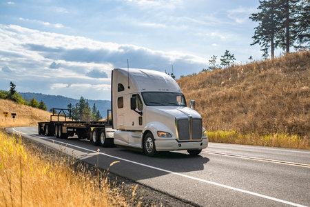 Industrial white long haul big rig high extended cab semi truck tractor transporting empty flat bed semi trailer running on the narrow winding highway road to warehouse for the next loadの写真素材