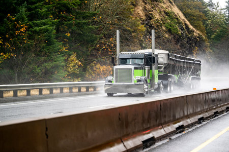Black and green classic big rig bonnet semi truck with chrome parts and high exhaust pipes transporting cargo in covered bulk semi trailer driving on the slippery wet road with rain and water dustの写真素材