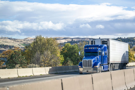 Industrial big rig blue semi truck tractor transporting frozen cargo in refrigerator semi trailer driving on elevated winding highway road to mountain pass in Californiaの写真素材