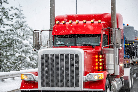 Industrial stylish bright red classic big rig semi truck with turned on auxiliary marker lights and chrome parts transporting cargo on flat bed semi trailer slowly skid on a winter snowy highway roadの写真素材