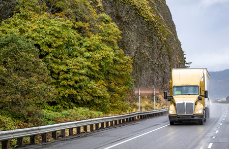Industrial standard big rig golden semi truck tractor transporting frozen cargo in reefer semi trailer driving on mountain winding highway road with rock cliff and fog and rain dust raised by wheelsの写真素材