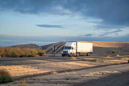 Industrial big rig white semi truck tractor with turned on headlight transporting commercial cargo in refrigerator semi trailer driving on the straight one way highway road at sunsetの写真素材