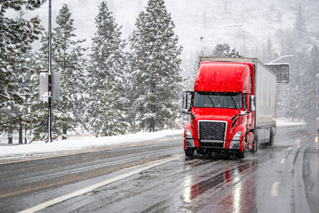 Red big rig commercial semi truck transporting cargo in dry van semi trailer running on the wet turning road with winter forest at snowing weather during a snow storm near Shasta Lake in Californiaの写真素材