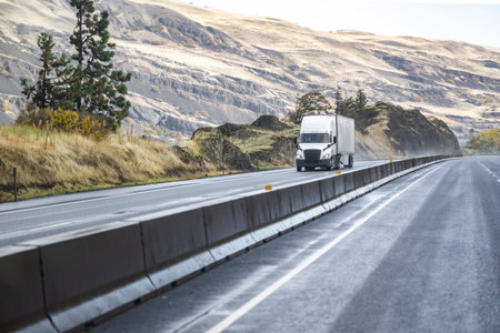 White industrial professional big rig long hauler semi truck with dry van semi trailer transporting commercial cargo running on the wet mountain highway road with rain dust from under the wheelsの写真素材