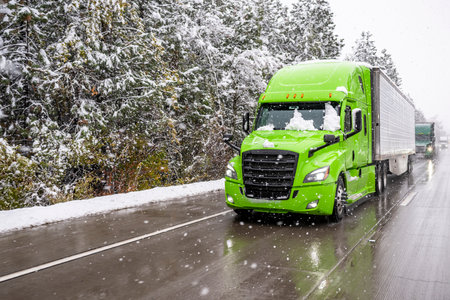 Bright green big rig semi truck transporting cargo in refrigerator semi trailer driving in front of convoy moving slowly on winter highway during a snow storm in the Lake Shasta region of Californiaの写真素材