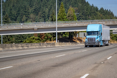 Industrial blue long haul big rig semi truck tractor transporting cargo in dry van semi trailer running on the straight wide highway road under the bridge and green forest on the backgroundの写真素材