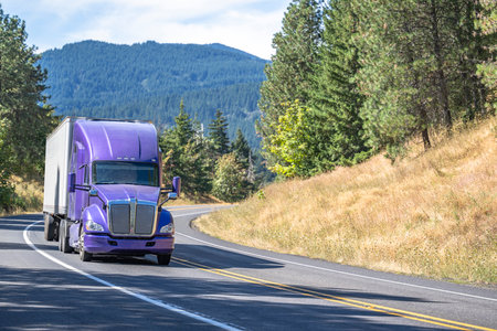 Commercial big rig purple semi truck tractor transporting frozen cargo in refrigerated semi trailer climbing uphill with load on the winding mountain road in Columbia Gorge areaの写真素材