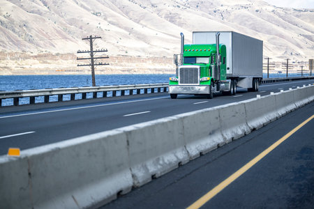 Industrial big rig green semi truck tractor with chrome grille transporting cargo in dry van semi trailer driving on divided highway road along the river with electric wind turbines in Columbia Gorgeの写真素材