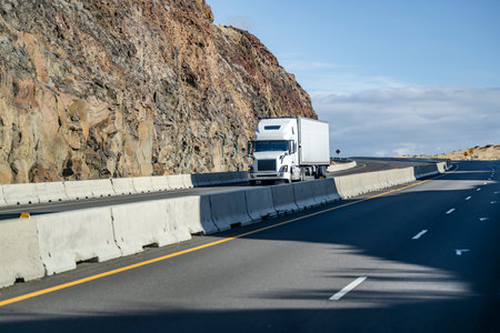 Industrial long hauler big rig white comfortable semi truck tractor transporting cargo in refrigerator semi trailer driving on the turning highway road in the mountain with rock wall on the sideの写真素材