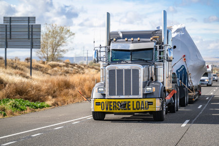 Industrial gray classic big rig semi truck tractor with oversize load sign and step down semi trailer transports oversized covered and fastened cargo driving on the highway road in Columbia Gorgeの写真素材