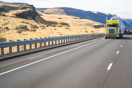 Industrial yellow classic big rig semi truck tractor with oversize load sign and step down semi trailer transports oversized covered and fastened cargo driving on the highway road in Columbia Gorgeの写真素材