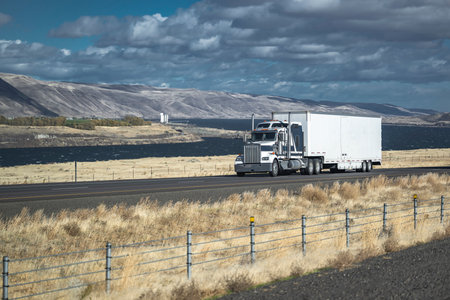 Industrial big rig gray semi truck tractor with chrome grille transporting cargo in dry van semi trailer driving on divided highway road along the river with electric wind turbines in Columbia Gorgeの写真素材