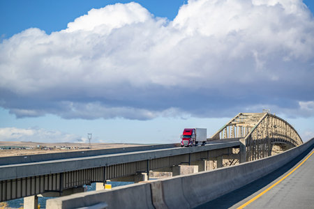 Industrial long hauler big rig red comfortable semi truck tractor transporting cargo in refrigerator semi trailer driving on the overpass road with truss bridge across the river at sunny dayの写真素材