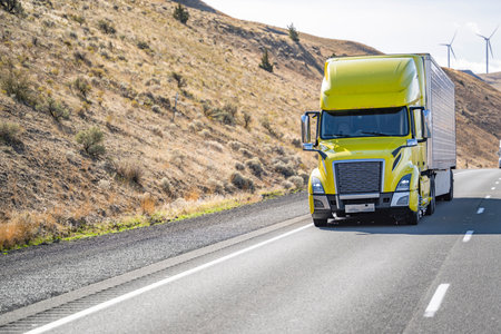 Industrial long hauler big rig yellow comfortable semi truck tractor transporting cargo in refrigerator semi trailer driving uphill on the divided highway road surrounded by hills in Washingtonの写真素材