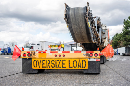 Industrial powerful big rig classic semi truck transporting oversized equipment on step down semi trailer with oversize load sign on the back standing for the rest on the truck stop parking lotの写真素材