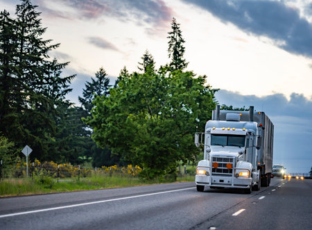 Long hauler big rig classic white semi truck tractor with turned on headlights and extended cab for truck driver rest transporting cargo in dry van semi trailer driving on the road at twilightの写真素材
