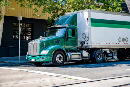 Local hauler powerful day cab big rig green semi truck tractor with roof spoiler transporting commercial cargo in dry van semi trailer make delivery of the cargo standing on the city streetの素材 [FY310207976910]