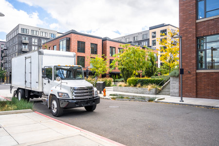 Industrial commercial compact middle duty rig white day cab semi truck tractor transporting cargo in box trailer standing on the urban city street with multilevel apartment buildingsの写真素材