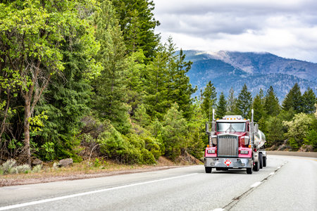 Industrial burgundy classic day cab big rig bonnet semi truck with chrome accessories transporting fuel in tank semi trailer driving on the mountain highway road with green forest on the sidesの写真素材