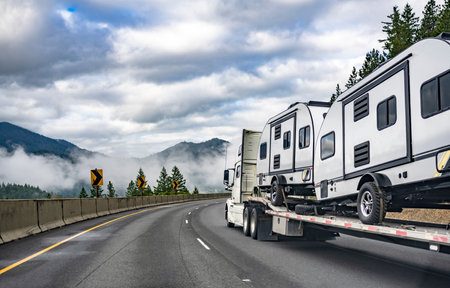 Industrial standard commercial big rig white semi truck tractor transporting campers trucks on the step down semi trailer driving on the winding highway road with fog on the forest and mountainの写真素材
