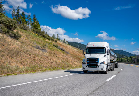 Industrial white low cab profile big rig bonnet semi truck with chrome accessories transporting fuel in tank semi trailer driving on the mountain highway road with green forest on the sidesの写真素材