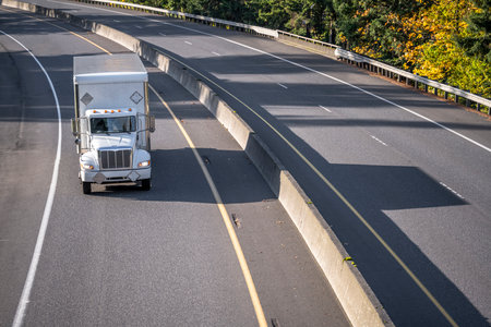Industrial standard commercial compact middle duty rig white day cab semi truck tractor transporting cargo in box trailer driving on the turning highway road at sunny day making local deliveriesの写真素材