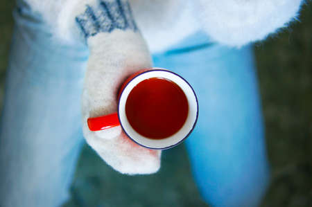 Red mug in one hand in mittens top view. Jeans and white sweaterの写真素材