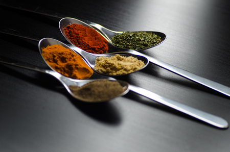 Spices colored in silver spoons, placed on a black wooden table. Pepper, pepper, salt, basil, mustardの写真素材