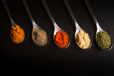 Spices colored in silver spoons, placed on a black wooden table. Pepper, pepper, salt, basil, mustardの写真素材