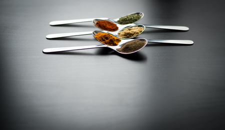 Spices colored in silver spoons, placed on a black wooden table. Pepper, pepper, salt, basil, mustardの写真素材