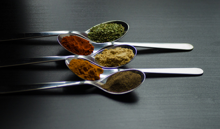 Spices colored in silver spoons, placed on a black wooden table. Pepper, pepper, salt, basil, mustardの写真素材
