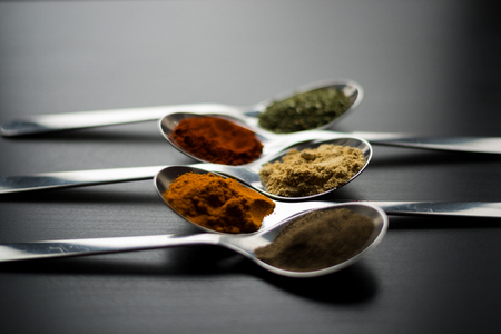 Spices colored in silver spoons, placed on a black wooden table. Pepper, pepper, salt, basil, mustardの写真素材