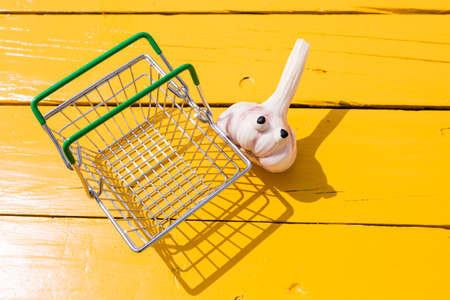 Funny garlic with eyes near the small supermarket basket on the yellow background. Vegetarian concept, copy spaceの写真素材