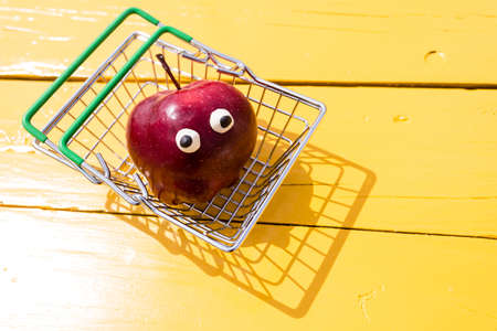Funny red apple with eyes in the small supermarket basket on the yellow background. Vegetarian concept, copy spaceの写真素材