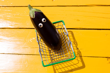 Funny purple eggplant with eyes in the small supermarket basket on the yellow background. Vegetarian concept, copy spaceの写真素材